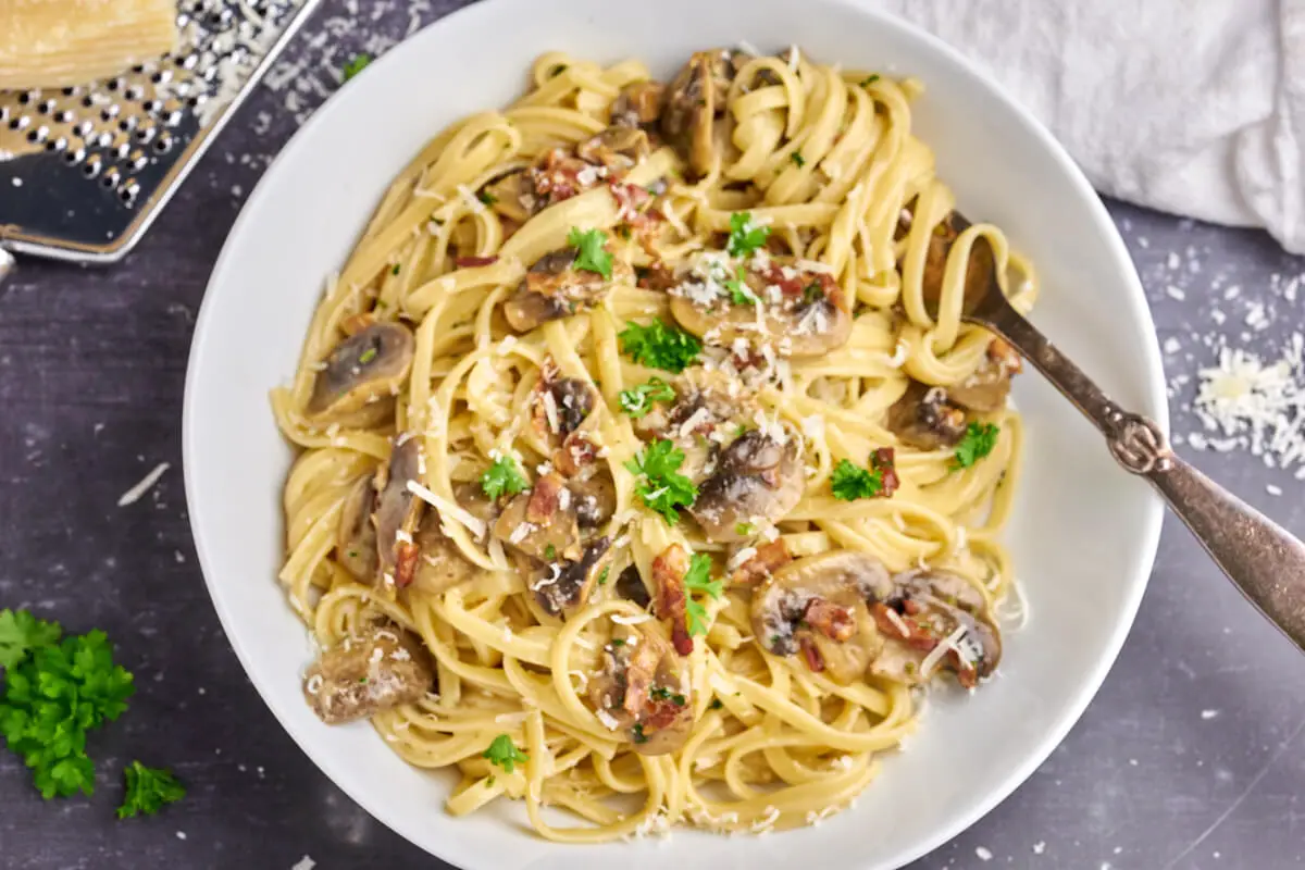 pasta med champignon, fløde og bacon i hvid skål med parmesan og persille på