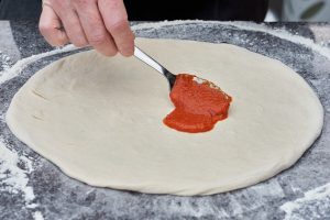 hjemmelavet italiensk pizzadej med pizzasauce der bliver smurt på med ske.