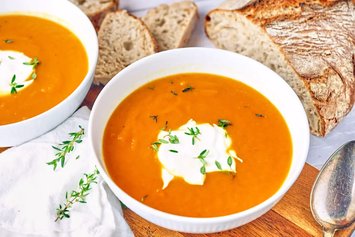 To hvide skåle med orange gulerodssuppe med creme fraiche og timian samt frisk brød