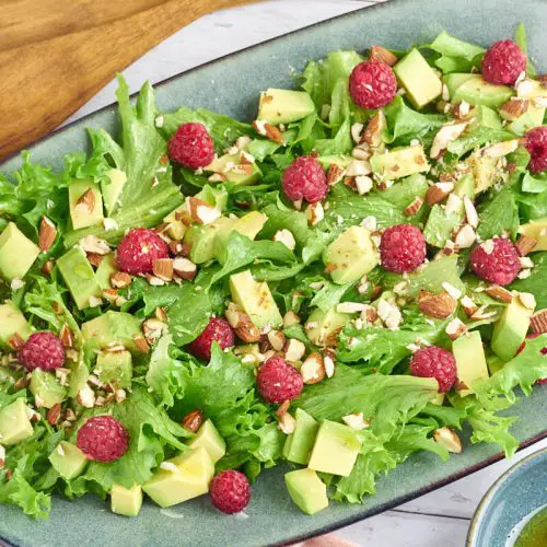 grøn salat med avocado og hindbær på ovalt fad med dressing og salatbestik