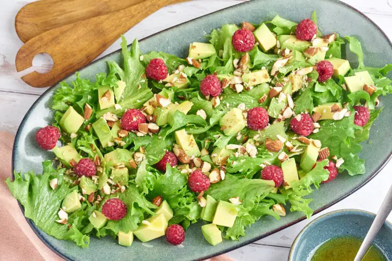 grøn salat med avocado og hindbær på ovalt fad med dressing og salatbestik
