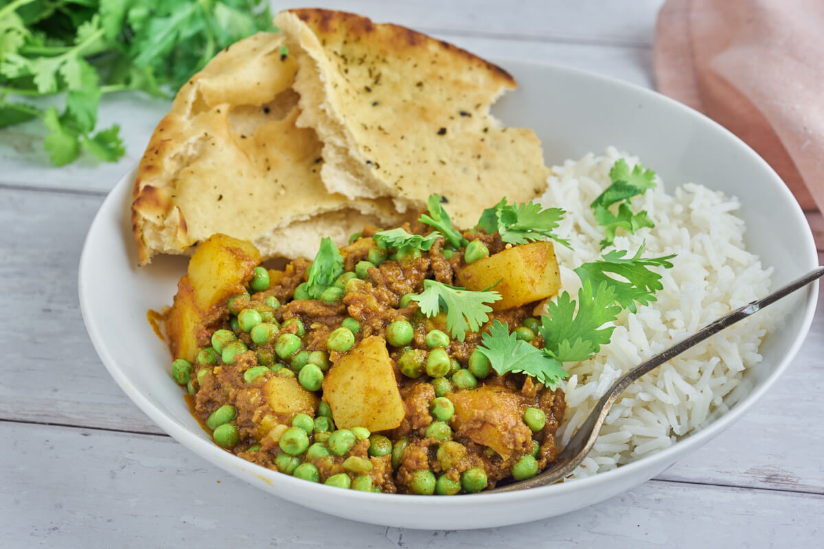 keema aloo med kartofler og ærter serveret i tallerken med kogte ris og naanbrød og drysset med frisk koriander