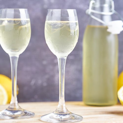 hjemmelavet limoncello i to glas med fod samt flaske og citroner i baggrunden