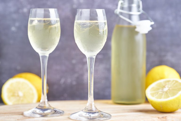 hjemmelavet limoncello i to glas med fod samt flaske og citroner i baggrunden