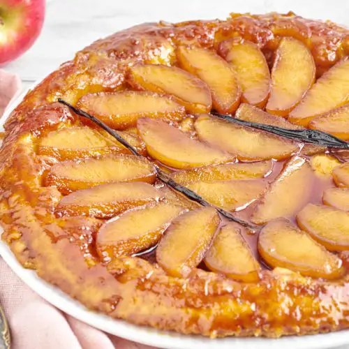 tarte tatin med æbler, karamel og vanilje på en hvid tallerken med kniv og friske æbler bagved