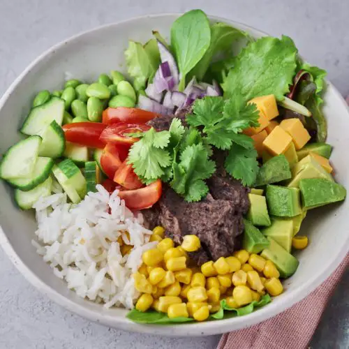 mexi bowl med sort bønnemos, hvide ris, majs, agurk, salat, tomat, cheddarost og andre grøntsager i skål