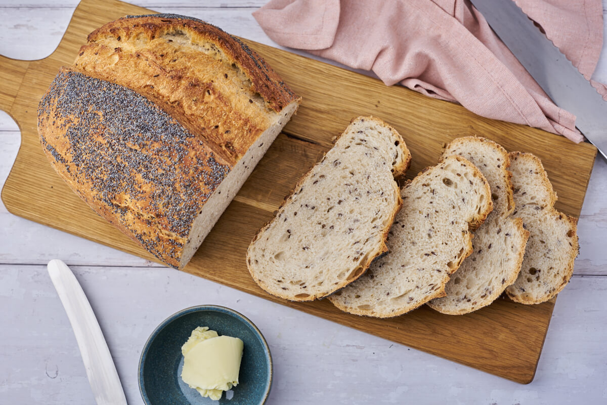 koldthævet skagensbrød med blå birkes på toppen og solsikkekerner i bunden samt hørfrø inden i på skærebræt med smør og kniv ved siden af