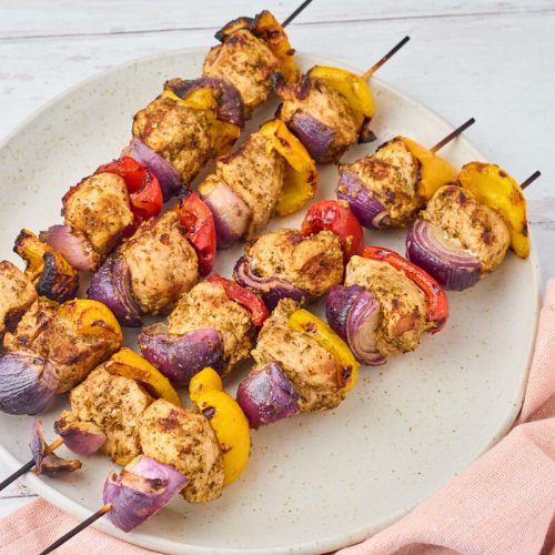 grillspyd med gyrosmarineret kylling og peberfrugt samt rødløg på serveringsfad.